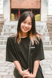 A young woman with black and light brown hair stands smiling in front of a stone staircase. She wears a black t-shirt and holds a small purse, with her hands intertwined. The background includes green foliage and part of a building with a tiled roof.