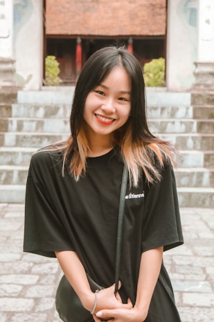 A young woman with black and light brown hair stands smiling in front of a stone staircase. She wears a black t-shirt and holds a small purse, with her hands intertwined. The background includes green foliage and part of a building with a tiled roof.