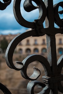 Intricate wrought iron scrollwork in the foreground with blurred architecture in the background. The sky appears cloudy, providing a contrast to the dark metal.