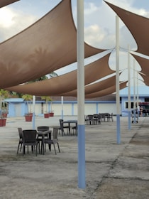 A modern outdoor patio area featuring tables and chairs beneath large, triangular fabric shades. The shades are suspended by tall poles and provide a contrast to the expansive concrete surface. Potted plants can be seen lining the edge of the area, adding a touch of greenery. In the background, there is a single-story building with blue and white walls, and a partially cloudy sky above.