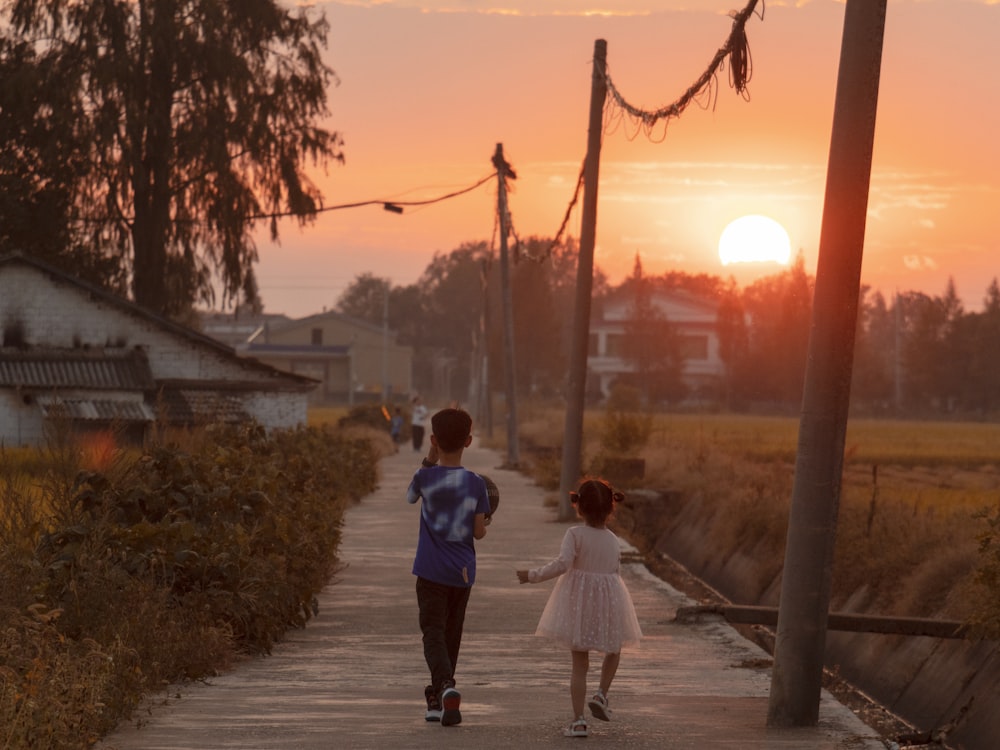 a couple of children walking down a sidewalk with a sunset in the background