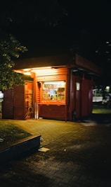 A small red kiosk or stand illuminated with yellow lights at night. It is situated on a paved sidewalk and surrounded by shadows and soft lighting from nearby street lights. The stand has a counter window, and some items are seen inside.