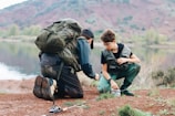 Two people are crouched by a lake, wearing outdoor gear and backpacks. They seem to be involved in a fishing activity, with a fishing rod placed on the ground nearby. The background features a calm lake and mountainous terrain with greenery.