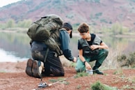 Two people are crouched by a lake, wearing outdoor gear and backpacks. They seem to be involved in a fishing activity, with a fishing rod placed on the ground nearby. The background features a calm lake and mountainous terrain with greenery.