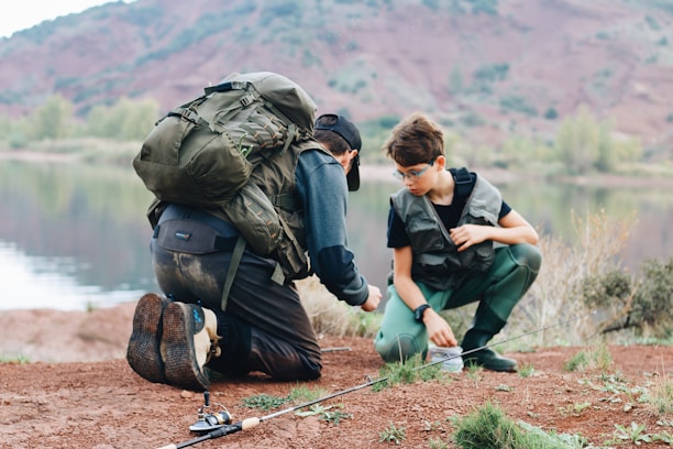 Two people are crouched by a lake, wearing outdoor gear and backpacks. They seem to be involved in a fishing activity, with a fishing rod placed on the ground nearby. The background features a calm lake and mountainous terrain with greenery.