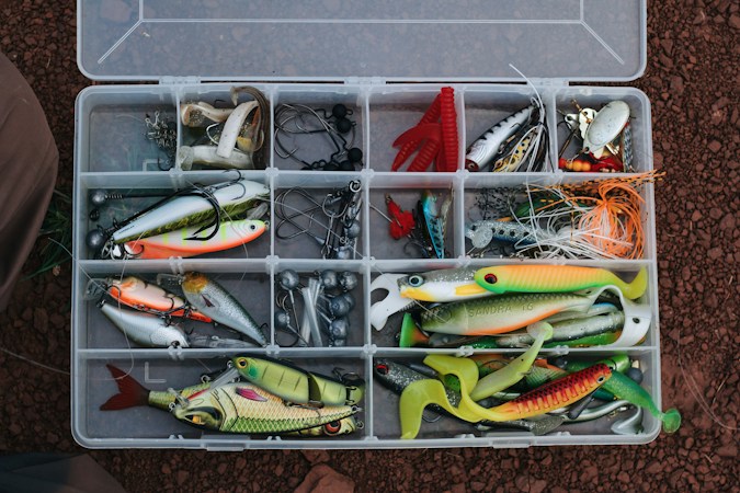 Open clear plastic tackle tray seen from above, its compartments filled with soft swimbaits, hard lures, jig heads and hooks, on red earth