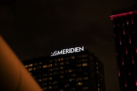 A nighttime cityscape featuring a building with the illuminated sign 'Le Meridien'. The sky is dark and overcast, and the building has some windows lit up. Another building with vertical pink lights is partially visible on the right side.