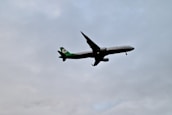 A commercial airplane is flying at a low altitude against a cloudy sky. The aircraft has a green tail and logo, while the rest of its body is predominantly white.