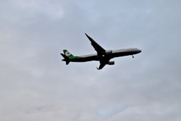 A commercial airplane is flying at a low altitude against a cloudy sky. The aircraft has a green tail and logo, while the rest of its body is predominantly white.