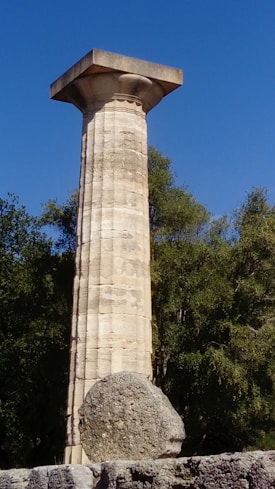 A tall, ancient Greek column stands prominently against a clear blue sky. The column features classic Doric design with fluted sides and a simple capital. In front of the column lies a large, spherical stone. Lush green trees surround the base of the column, providing a natural backdrop.