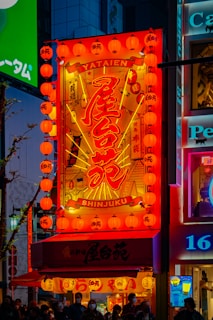 A bright and colorful illuminated sign with Japanese characters and the words 'YATAIEN SHINJUKU'. The sign is surrounded by orange lanterns that emit a warm glow, creating a vibrant and lively atmosphere typical of an urban street at night.