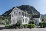 A quaint white house with intricate decorative trim stands against the backdrop of a majestic, green-covered mountain. The structure appears to be a two-story building with large windows and a classic roof. In the foreground, there are potted plants and two bicycles leaning against the fence. A small chalkboard sign sits near some outdoor seating, suggesting a welcoming and relaxed atmosphere.