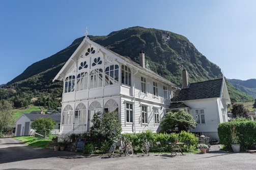 A quaint white house with intricate decorative trim stands against the backdrop of a majestic, green-covered mountain. The structure appears to be a two-story building with large windows and a classic roof. In the foreground, there are potted plants and two bicycles leaning against the fence. A small chalkboard sign sits near some outdoor seating, suggesting a welcoming and relaxed atmosphere.