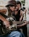 A man with long hair and a beard is playing an acoustic guitar adorned with floral decals. He is wearing a cowboy hat and a sleeveless shirt. Another person, with a slicked-back hairstyle and wearing a dark sleeveless shirt, leans in from behind, holding a cigarette. The scene appears to be outdoors, possibly in a rustic or street setting.