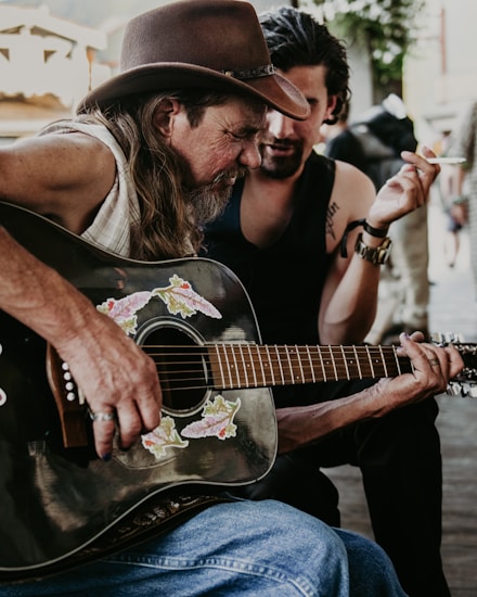A man with long hair and a beard is playing an acoustic guitar adorned with floral decals. He is wearing a cowboy hat and a sleeveless shirt. Another person, with a slicked-back hairstyle and wearing a dark sleeveless shirt, leans in from behind, holding a cigarette. The scene appears to be outdoors, possibly in a rustic or street setting.