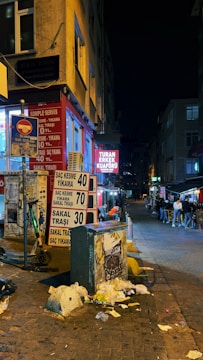 A nighttime street scene in an urban area. Illuminated shop signs and streetlights line the street. A red and white signage prominently displays prices for haircuts and beard trims. The building walls and the street appear slightly worn. People are walking and socializing in the background. Trash and debris are visible on the ground.