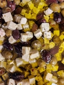 A mixture of olives and cubes of white cheese, possibly feta, is immersed in olive oil. The olives vary in color, including green and purple hues. The cheese is seasoned with herbs and is evenly distributed among the olives.