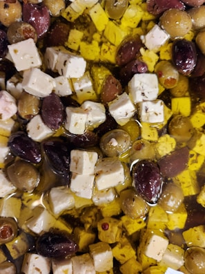 A mixture of olives and cubes of white cheese, possibly feta, is immersed in olive oil. The olives vary in color, including green and purple hues. The cheese is seasoned with herbs and is evenly distributed among the olives.