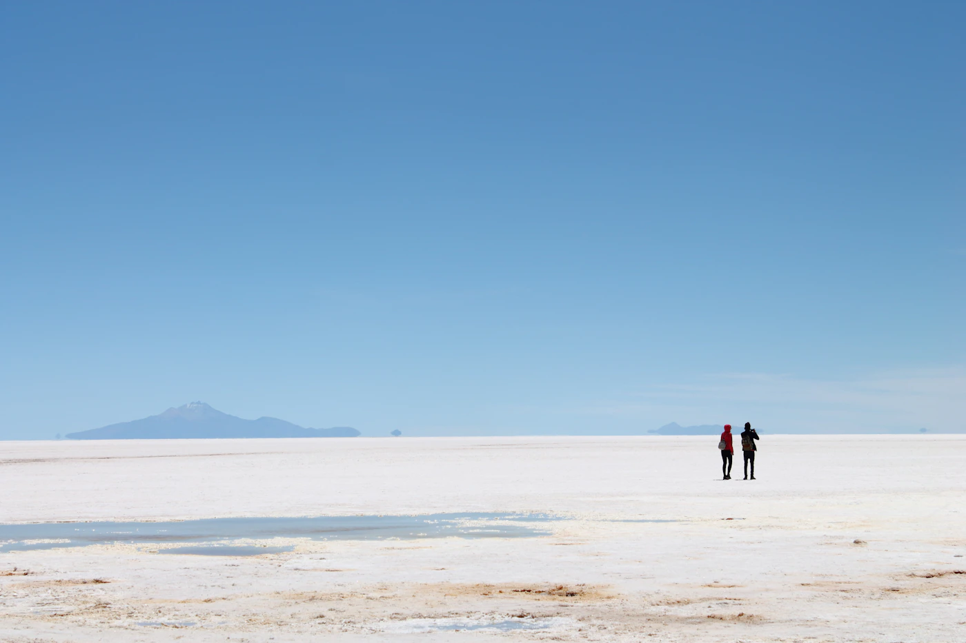Couple in elopement ceremony attire on the crest of a sand dune with the vast desert landscape extending to the horizon in all directions and the warm evening light illuminating them from the side