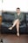 A woman is sitting on a black leather sofa, wearing a green dress and black high-heeled sandals. She has an arm resting on the back of the sofa and her legs crossed, exuding a relaxed and confident posture. A bottle of wine is placed on the wooden floor in front of the sofa.