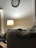 A cozy room with a lighted lamp on a small table near a wall adorned with a clock. A dog is comfortably nestled on a couch, partially covered by a plaid blanket.