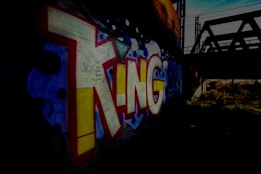 Large, colorful graffiti art spelling 'KING' in bold letters on a brick wall, set in a dimly lit underpass or tunnel with a nearby bridge structure visible in silhouette.