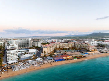 Aerial view of a beachside resort area with expansive sandy shores and clear turquoise waters. Numerous buildings, including hotels and resorts, line the coastline. Beach umbrellas and lounge chairs are neatly arranged on the sand, and the area is bustling with activity. The landscape extends into the background with hills and a mountain range in the distance, under a partly cloudy sky.