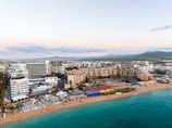 Aerial view of a beachside resort area with expansive sandy shores and clear turquoise waters. Numerous buildings, including hotels and resorts, line the coastline. Beach umbrellas and lounge chairs are neatly arranged on the sand, and the area is bustling with activity. The landscape extends into the background with hills and a mountain range in the distance, under a partly cloudy sky.