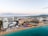 Aerial view of a beachside resort area with expansive sandy shores and clear turquoise waters. Numerous buildings, including hotels and resorts, line the coastline. Beach umbrellas and lounge chairs are neatly arranged on the sand, and the area is bustling with activity. The landscape extends into the background with hills and a mountain range in the distance, under a partly cloudy sky.