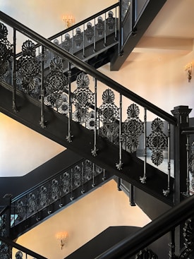 An ornate, intricately designed black staircase with detailed patterns on the railing. The staircase features multiple flights and is set against a light-colored wall. Elegant wall-mounted candle-like fixtures provide soft lighting in the background.