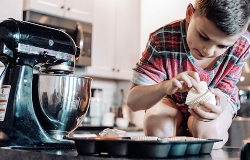 A young boy is focused on placing cupcake liners into a baking tray. He is wearing a plaid shirt and is near a large black kitchen mixer on a countertop. The kitchen background includes cabinets and various kitchen items.