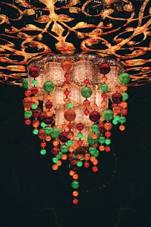A lavish and ornate chandelier adorned with hanging colorful glass orbs in shades of red, green, and gold. The chandelier features intricate designs and patterns with sparkling crystals, creating a luxurious and festive appearance.