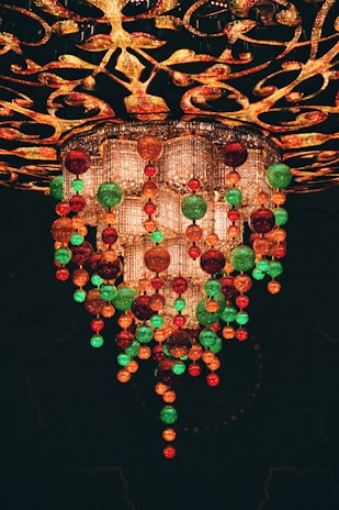 A lavish and ornate chandelier adorned with hanging colorful glass orbs in shades of red, green, and gold. The chandelier features intricate designs and patterns with sparkling crystals, creating a luxurious and festive appearance.