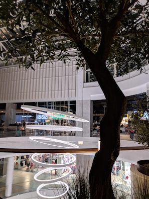 A large, modern shopping mall interior features multiple floors with shops and a food court. A series of spiral-shaped, illuminated rings hang from the ceiling as decorative elements. A tree with lush green leaves stands in the foreground, adding a touch of nature to the setting.