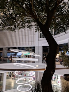 A large, modern shopping mall interior features multiple floors with shops and a food court. A series of spiral-shaped, illuminated rings hang from the ceiling as decorative elements. A tree with lush green leaves stands in the foreground, adding a touch of nature to the setting.
