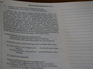 A close-up view of an open book showing text from Ecclesiastes 4:1. The text is typed in a serif font, and the page features numbered verses with a visible heading. The content discusses the human condition and philosophical reflections. The lighting is soft, casting slight shadows.
