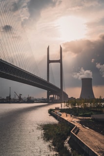 A large cable-stayed bridge stretches across a wide river with a cooling tower emitting steam in the background. The scene is partially illuminated by the sun peeking through clouds, casting a warm glow. The waterfront area has a pathway lined with trees and a couple of parked cars.