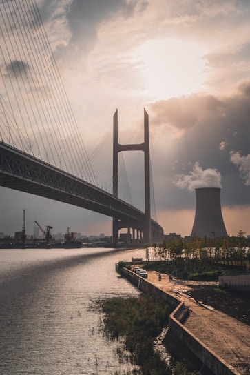 A large cable-stayed bridge stretches across a wide river with a cooling tower emitting steam in the background. The scene is partially illuminated by the sun peeking through clouds, casting a warm glow. The waterfront area has a pathway lined with trees and a couple of parked cars.