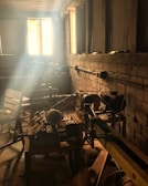 A rustic workshop setting with sunlight streaming through a window, illuminating dust particles in the air. Various woodworking tools, such as a lathe, are visible on the workbench, and wooden chairs are scattered around the room. The walls are unfinished brick, adding to the workshop's vintage feel.