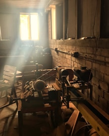 A rustic workshop setting with sunlight streaming through a window, illuminating dust particles in the air. Various woodworking tools, such as a lathe, are visible on the workbench, and wooden chairs are scattered around the room. The walls are unfinished brick, adding to the workshop's vintage feel.