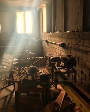A rustic workshop setting with sunlight streaming through a window, illuminating dust particles in the air. Various woodworking tools, such as a lathe, are visible on the workbench, and wooden chairs are scattered around the room. The walls are unfinished brick, adding to the workshop's vintage feel.