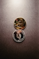 Two Ethereum coins, one gold and one silver, are placed on a textured surface. The coins feature the Ethereum logo and inscriptions related to decentralized platforms and smart contracts.