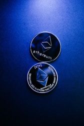 Two Ethereum coins with a metallic surface lie against a dark blue background. The top coin displays the Ethereum logo and text, while the bottom coin highlights the phrases 'decentralized platform' and 'smart contracts'.