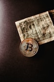 A golden Bitcoin coin is placed next to a crumpled US dollar bill on a dark surface, symbolizing the contrast between cryptocurrency and traditional currency.