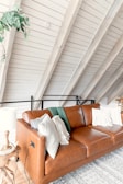 A cozy living space featuring a brown leather couch with multiple decorative pillows and a throw blanket. The couch is positioned against a white paneled wall with exposed beams. There's a small wooden side table beside the couch with a lamp on it. A green plant hangs from the ceiling above the couch. The room has an inviting and comfortable atmosphere with a touch of rustic charm.