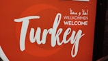 A sign featuring the word 'Turkey' in large, artistic script with the word 'Welcome' underneath in multiple languages, including English, German, and Arabic. The background is predominantly red with a circular design element.