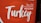 A sign featuring the word 'Turkey' in large, artistic script with the word 'Welcome' underneath in multiple languages, including English, German, and Arabic. The background is predominantly red with a circular design element.