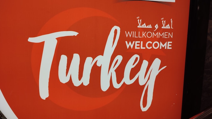 A sign featuring the word 'Turkey' in large, artistic script with the word 'Welcome' underneath in multiple languages, including English, German, and Arabic. The background is predominantly red with a circular design element.