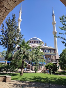 A large mosque with two tall minarets and a central dome, surrounded by lush green trees under a clear blue sky. The architectural design is intricate, with multiple arched windows and decorative elements. A neatly maintained garden with grass and shrubs enhances the foreground.