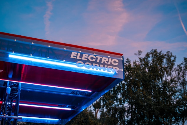 A modern structure with the words 'Electric Corner' prominently displayed in bright lights. The canopy appears metallic with a combination of red and blue accent lighting. The background shows a clear evening sky with some clouds and trees.